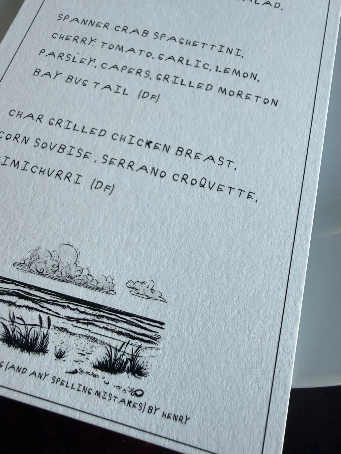 Close-up of a wedding menu showing a coastal illustration and a handwriting credit line