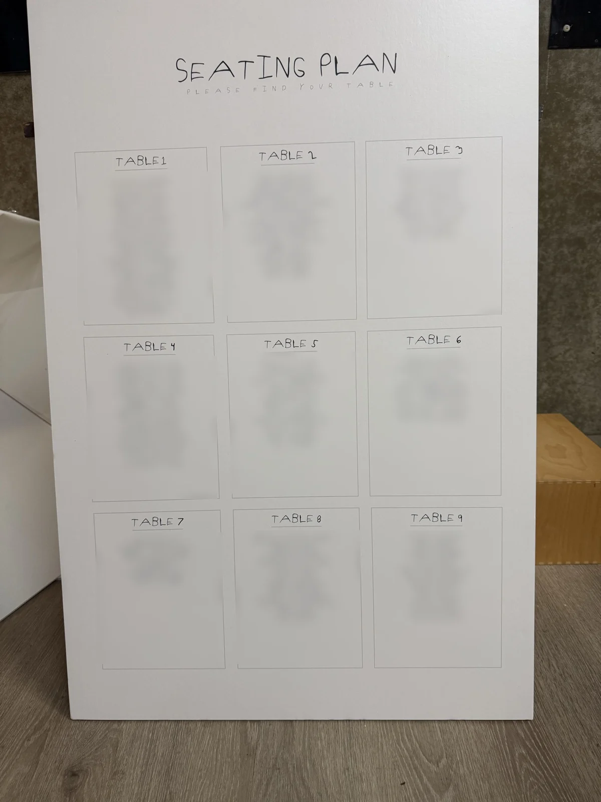 Wedding seating plan board with nine tables in handwritten lettering, guest names blurred for privacy
